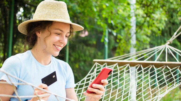 A woman in a straw hat holding a credit card and her phone booking travel.